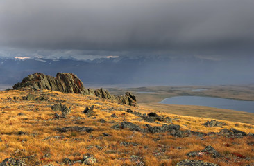 Colorful rock and dry yellow grass in the background of highland steppe lake with snow covered mountains under a stormy cloudy dramatic rain sky Plateau Ukok, Altai, Siberia, Russia