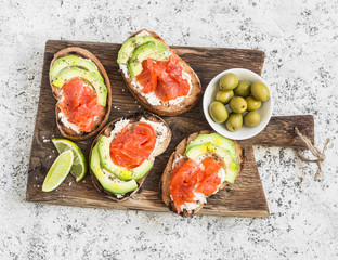 Delicious appetizers - cream cheese, smoked salmon and avocado sandwiches and olives on a wooden board. On a light background, top view