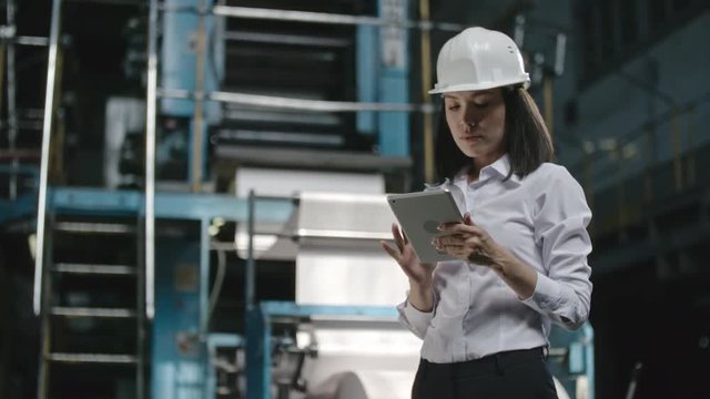 Female executive engineer in hard hat standing in factory workshop looking up, then writing something down on her tablet