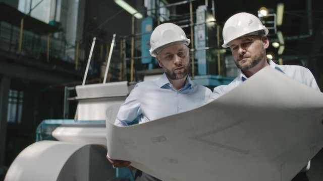 Male architects in hard hats standing in factory workshop holding layout plan and discussing it