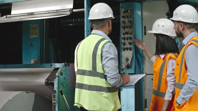 Male and female engineer standing before industrial paper machine and describing its work to inspector in safety vest and hard hat making notes