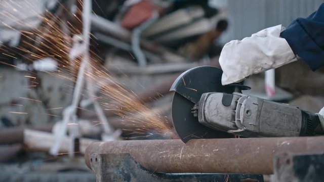 Hands of male worker in protective gloves holding grinder tool and cutting metal pipe with sparks flying from it