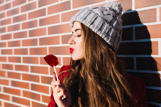 Closeup portrait attractive brunette girl with long hair on wall background outside. She wears knitted hat,  keeps eyes closed, sends kiss to lollipop red lips