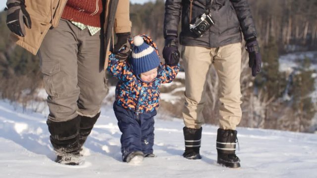 Tracking of man and woman leading toddler by hands and helping him walk through deep snow in winter