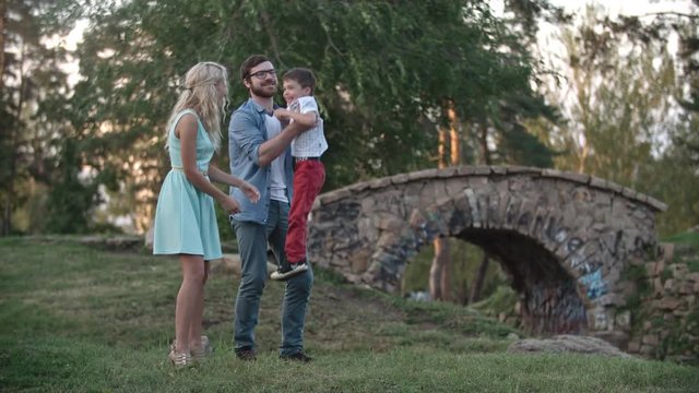 Parents spending time with their son in the park, dad carrying son in his arms and mom joyfully tickling him 