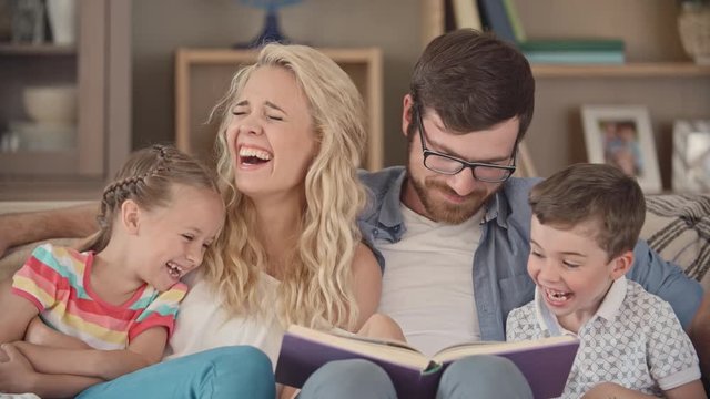 Parents and their little kids sitting together on couch and looking at the book, mom pointing at something funny, kids laughing happily
