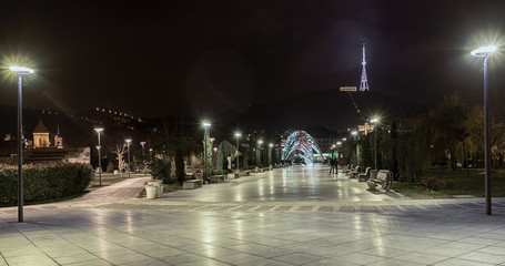 Fototapeta premium Georgia, Tbilisi - November 23, 2016. Panorama of the night city. Recreation Park Rijk, TV tower, pedestrian bridge Peace empty benches and light on the lampposts.