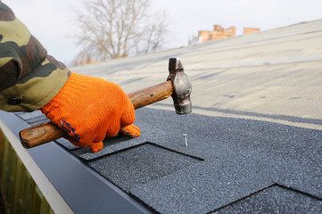 Builder Building Roof. Worker Hammer in Nails on the Roof. Roofer is hammering a Nail in the Roof Shingles. Construction Nails Vapor barrier and Waterproofing. Unfinished roof.