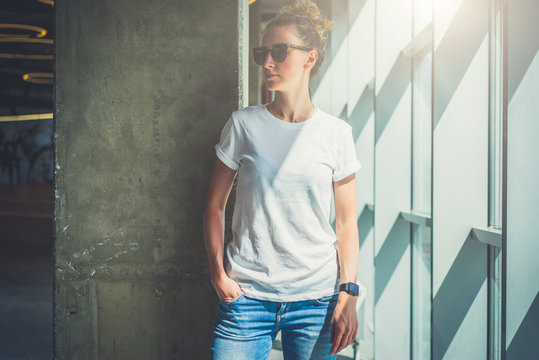 Girl with blonde hair in sunglasses,white T-shirt and blue jeans is standing in room on background of concrete column next to window.  Bright sunlight. Mock up.