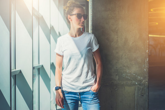 Girl with blonde hair in sunglasses,white T-shirt and blue jeans is standing in room on background of concrete column next to window. On hand of young woman digital gadget - smartwatch. Mock up.