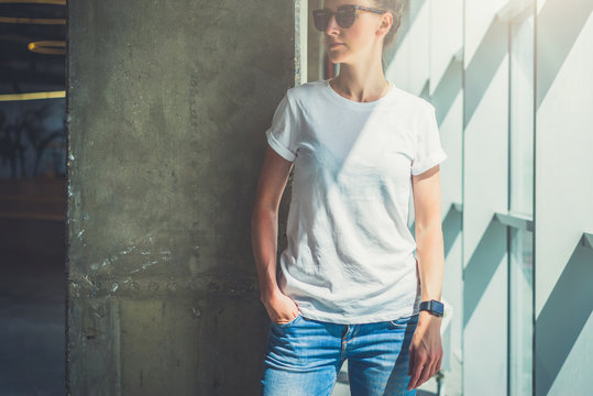 Girl in sunglasses,white T-shirt and blue jeans standing with her hand in pocket jeans in a room against concrete wall next to window. On hand of young woman digital gadget - smartwatch. Mock up.
