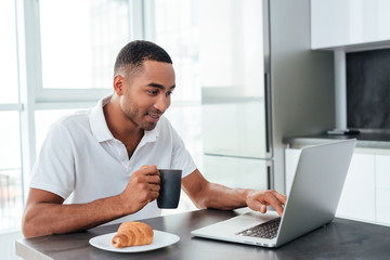 Smiling african man drinking coffee and using laptop on kitchen