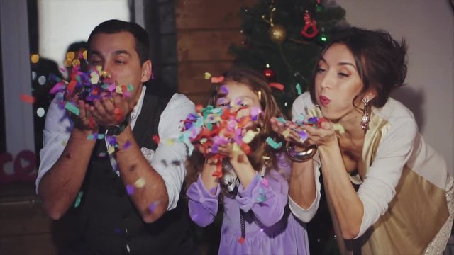 Slow motion shot of happy mom, dad and daughter blowing colorful confetti at home. Family celebration of Christmas and New Year