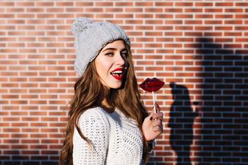 Portrait pretty brunette girl in white sweater and knitted hat on wall background outside. She holds lollipop red lips, smiling  to camera