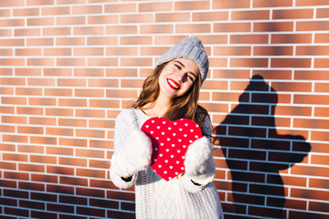 Portrait pretty girl with long hair in knitted hat, warm sweater on wall background outside. She holds red big heart in gloves, smiling to camera