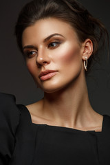 Fototapeta premium Portrait of a beautiful, self-sufficient and confident young woman with collected hair in the elegant black dress with makeup, dark background, studio photo