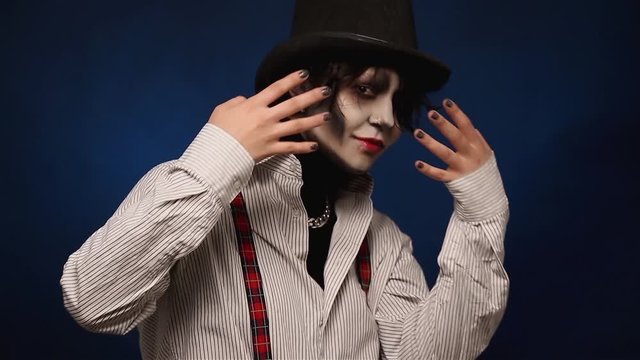 Portrait of beautiful girl with Halloween makeup closing her eyes. Attractive young woman in white shirt and with black hat on hiding from camera in studio.