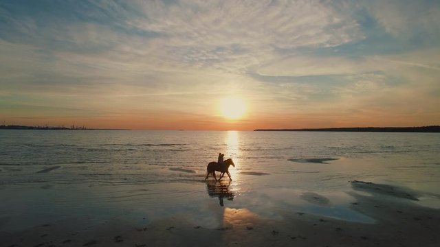 Two GIrls are Riding Horses on a Beach. Horses Walk on Water. Beautiful Sunset is Seen in this Aerial Shot.