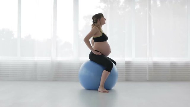 A young pregnant woman is jumping on a blue fitball in the fitness room.