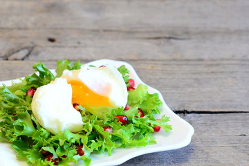 Poached egg, lettuce leaves with pomegranate seeds and olive oil. Healthy salad on a plate on wooden background with copy space for text. Healthy diet egg recipe. Closeup