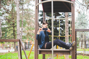 Obraz premium Young beautiful short brown hair woman sitting in a playgroung holding a smart phone taking photos - technology, social network, communication concept