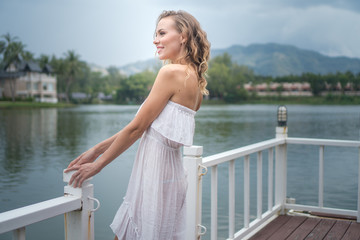 Fototapeta na wymiar Pretty blond woman in sexy white summer dress standing on the pier and smiling against beautiful lake and tropical resort background. Beautiful female enjoying her holiday, vacation concept