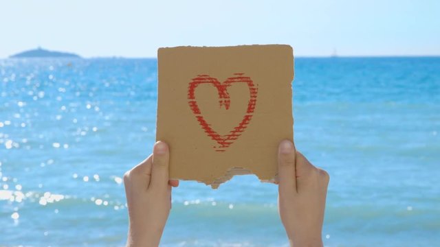 Sequence of different shots showing a hand drawn heart on a piece of cardboard, held by a young woman standing on the beach in front of the beautiful blue sea - ProRes