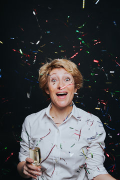 Excited middle aged woman with glass of champagne laughing in flying colorful confetti isolated on black.