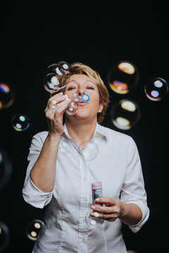 Middle aged woman in casual outfit blowing soap bubbles isolated on black