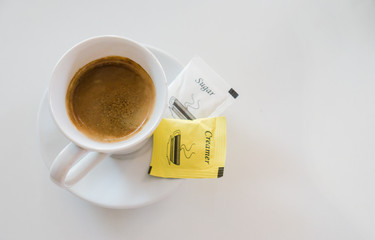 Hot black coffee in white cup with cream and sugar in packet on white table 