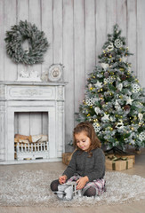 Pretty little girl sits on a floor and plays with wooden toy near a fireplace in Christmas at home