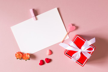 Valentine day composition: red gift box with bow, stationery / photo / postcard template with clamp, small hearts, candy and spring flowers on light pink background. Top view.