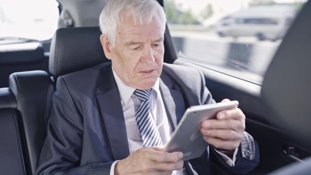 Senior businessman communicating via tablet and checking his wristwatch while sitting in fast moving car