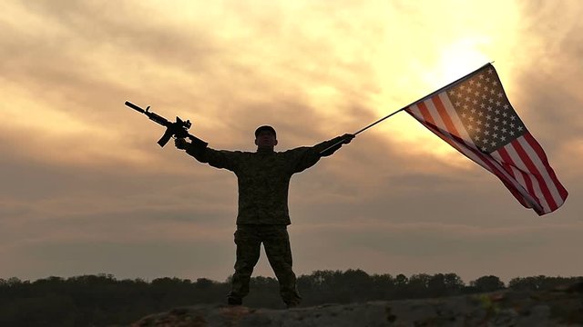 
Soldier stand with American flag and rifle against gloomy orange  sky. Slow Motion
