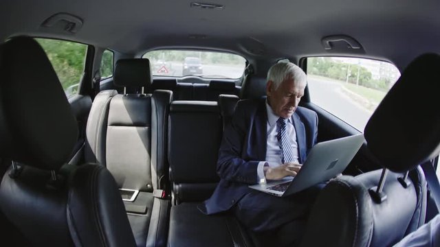 Fish eye shot of senior businessman sitting in moving car talking on phone and working on his laptop