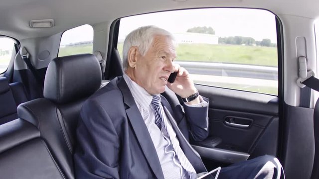 Senior businessman with grey hair sitting in moving car and arguing with somebody over mobile phone
