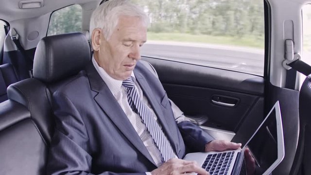 Senior politician in suit sitting in moving car and typing on laptop