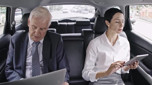 High angle shot from inside of car of senior businessman with laptop working in moving car next to his female colleague communicating with partners on tablet