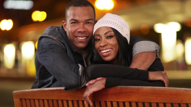 Portrait Of Black Man And Woman Looking Over Back Of Bench On City Street At Night