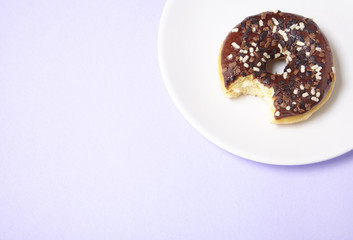 An iced chocolate sprinkle ring donut with bite mark, on a pastel purple background with blank space at side