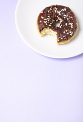 An iced chocolate sprinkle ring donut with bite mark, on a pastel purple background with blank space below