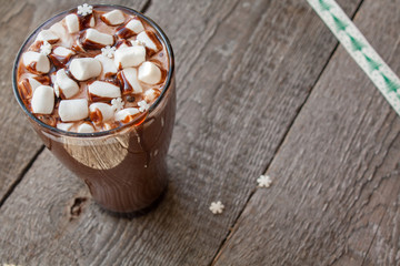 Winter hot cocoa with marshmallows, chocolate syrup, sugar snowflakes in a tall glass on a wooden background.