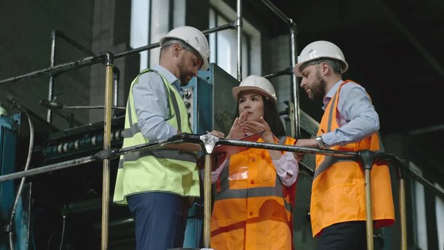 Low angle shot of male and female engineer in hard hats and reflective safety vests standing near industrial machinery and cheerfully talking to inspector with clipboard 