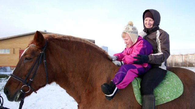 Hippotherapy for kid with cerebral palsy syndrome at winter cold day - contact kids therapy and rehabilitation horse-riding club - young girl and her mother rides on horse, pats horseback