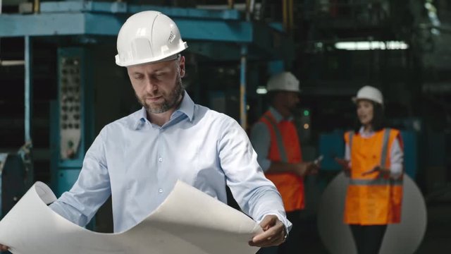 PAN of male engineer in formalwear and hard hat standing at factory inspecting blueprints, then looking at camera and smiling 