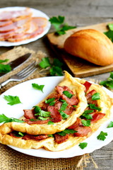 Homemade bacon omelette. Delicious omelette stuffed with bacon and parsley on a plate, bacon slices, bread, fork, knife, cutting board on old wooden table. Rustic style breakfast concept. Closeup