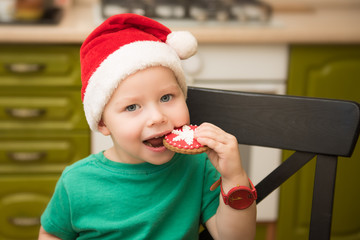 The boy eats the cookie in the hat of Santa Claus sitting in the kitchen