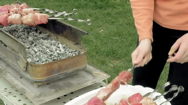 Girl puts barbecue bbq skewers with red meat on brazier with hot coals embers. Outdoors picnic concept.