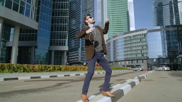 Handsome man listening to music in smartphone headphones and dancing near skyscrapers