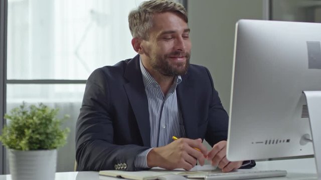 Handsome bearded businessman talking with smile while video calling on his computer in the office